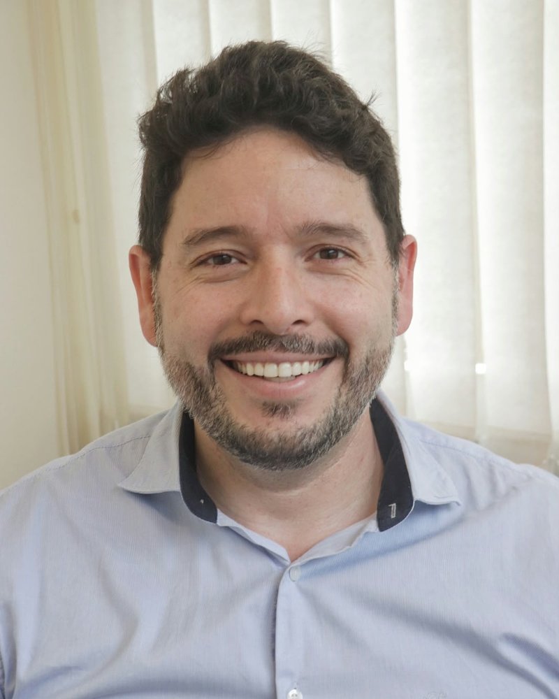 Retrato de Gustavo Lowenberg Scalco, psicólogo e psicanalista, sorrindo, de camisa azul-clara
